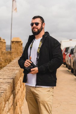 Genç sakallı, güneş gözlüklü, köprüde kamerası olan bir turistin dikey fotoğrafı.