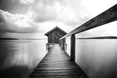 Almanya 'nın Bavyera kentindeki bulutlu gökyüzüne karşı Ammersee Gölü üzerindeki bir kayıkhanenin gri ölçekli görüntüsü.