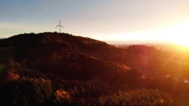 Orman ağaçları ve rüzgar türbiniyle kaplı tepede parlayan güneş batımının insansız hava aracı görüntüsü
