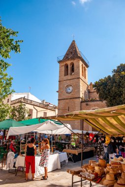 Son Servera 'da market tezgahları, alışveriş yapan insanlar ve arka planda kilise olan bir pazar.
