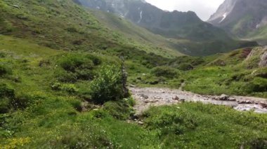 Yeşil dağların güzel manzarası, küçük bir su yolu ve çimlerin üzerinde oturan insanlar.