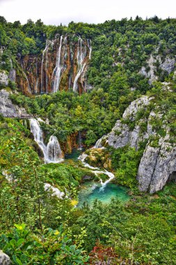 Yeşil ağaçlarla çevrili kayaların ve Plitvice göllerinin üzerinden akan şelalelerin manzarası.