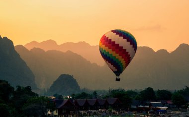 Sıcak hava balonu gün batımında bir dağ sırasının arka planına sahip bir kasabanın üzerinde uçuyor, Laos.
