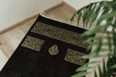 Köşe başında Kabe desenli siyah kadife bir seccadilin arka planında bir ev bitkisinin olduğu bir yer.