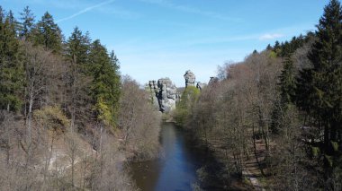 Arka planda çoğunlukla kuru ağaçlarla çevrili bir nehir manzarası ve dış taraftaki kumtaşı kaya oluşumu.