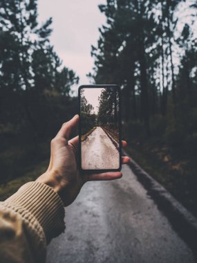 Akıllı telefonu tutan ve ormanın içinden geçen bir yolun fotoğrafını çeken bir erkeğin dikey görüntüsü.