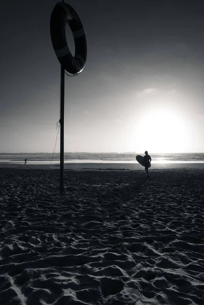 在海滩上一个救生圈附近的一个冲浪运动员的轮廓的垂直灰度照片 — 图库照片
