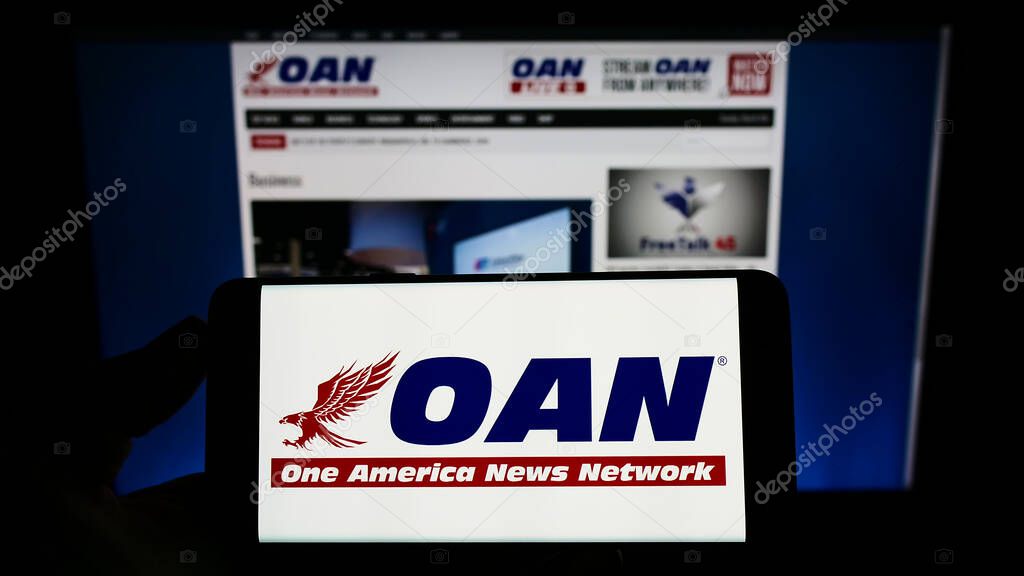 Person holding cellphone with logo of American media company One America News Network (OANN) on screen in front of website. Focus on phone display.