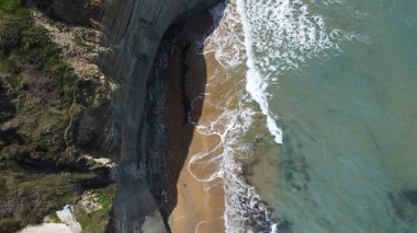 Uçurumların altında turkuaz deniz dalgaları tarafından yıkanan huzurlu kumlu bir sahil manzarası.