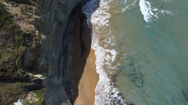 Uçurumların altında turkuaz deniz dalgalarıyla yıkanmış kumlu bir sahil manzarası.