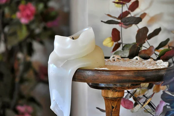 A white, pillar candle that had burned and melted down sets on the edge of a doilie covered, round, side table.