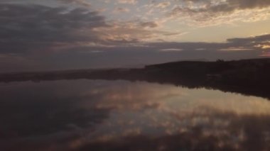 Gün batımında sahilde evleri ve ağaçları yansıtan bir deniz manzarası.