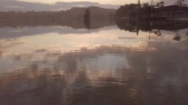 Gün batımında sahilde evleri ve ağaçları yansıtan bir deniz manzarası.