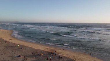 Güneşli bir günde kumlu bir sahilde okyanus dalgalarına karşı eğlenen insanların kuş bakışı görüntüsü.