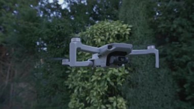DJI Mavic Mini insansız hava aracı yeşil ağaçların ve çalıların önünde uçuyor, soldan sağa dönüyor, görüntüden çıkıyor.