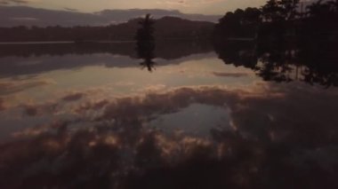 Gün batımında sahilde evleri ve ağaçları yansıtan bir deniz manzarası.