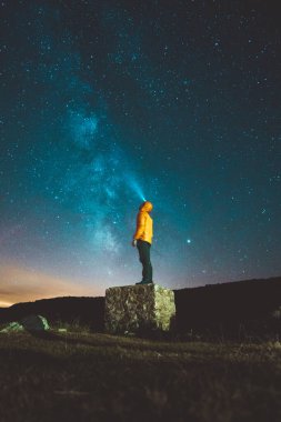 Sarı ceketli bir adamın dikey görüntüsü bir kayanın üzerinde duruyor ve Samanyolu 'nun güzel manzarasının tadını çıkarıyor.