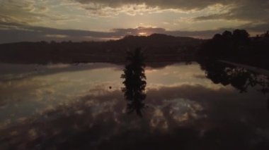 Gün batımında sahilde evleri ve ağaçları yansıtan bir deniz manzarası.