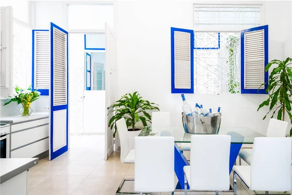 An interior home with white with blue furniture windows and green plants showing the dining area and kitchen
