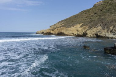 Mavi gökyüzünün altındaki dağlık sahil şeridinde dalgalı ve köpüklü bir suyun manzarası.