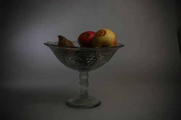 A decorative glass vase with rotten fruits on a gray background