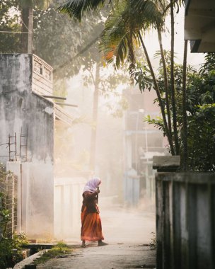 Güneşli bir sokakta kafa örtüsü takmış yalnız bir kadının dikey görüntüsü.