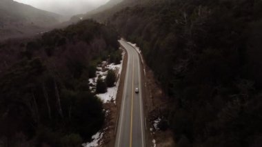 Patagonya 'da bir ormanın ortasındaki yolda giden bir arabanın yükselen hava görüntüsü.
