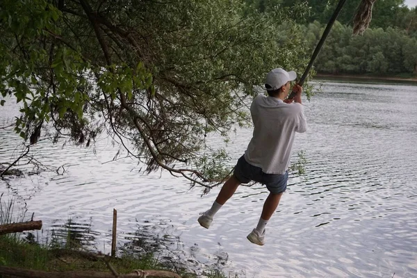 緑の木で湖の水の上にロープでスイング若い男性のバックショット — ストック写真