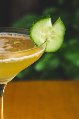 Yeşil bitkilere karşı salatalıkla süslenmiş bardaktaki bir kokteylin dikey yakın çekimi.