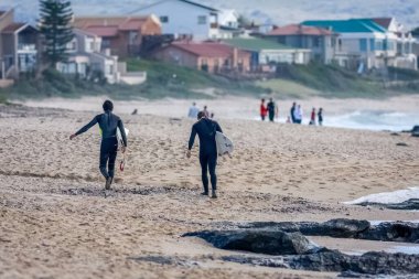 Jeffreys Bay, Güney Afrika 'da sörf tahtasıyla sahilde yürüyen dalgıç kıyafetli sörfçülerin arka planı.