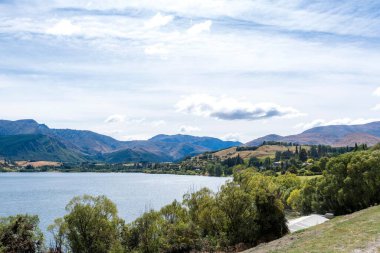 Parlak bulutlu bir gökyüzünün altında ağaçlar ve tepelerle çevrili bir su kütlesinin manzarası.