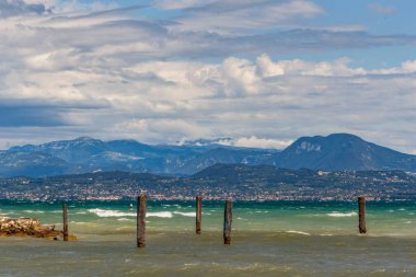 Su yüzeyindeki kazıklar bulutlu mavi gökyüzü ile tepelere karşı. Garda, İtalya.