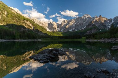 Güneşli bir günde yeşil ve karlı dağlarla çevrili bir göl manzarası.