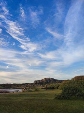 Bulutlu mavi gökyüzünün altındaki yeşil ağaçlara ve tepelere bakan bir gölün dikey görüntüsü.