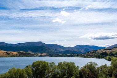 Parlak bulutlu bir gökyüzünün altında ağaçlar ve tepelerle çevrili bir su kütlesinin manzarası.