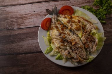 Tahta bir masada beyaz bir tabakta sunulan tuzla pişirilmiş çipura ve sarımsakla kaplı bir salata..