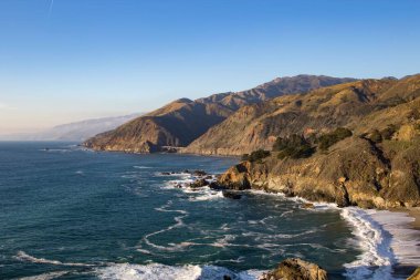 Kaliforniya 'da yeşilliklerle kaplı kıyı dağlarına çarpan okyanus dalgalarının hava görüntüsü.