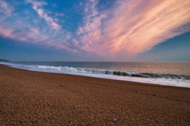 Gün batımında kumlu bir sahile vuran dalgaların manzarası çizgili bir gökyüzüyle