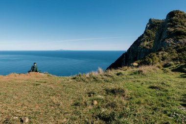 Mavi deniz ve gökyüzüne karşı yeşil uçurumun kenarında oturan adam. Madeira, Portekiz.