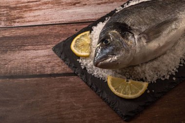 Taze deniz çipurasının yakın plan çekimi. Tahta bir masada siyah bir levha üzerinde limon dilimleri.