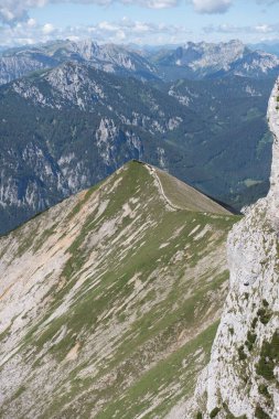 Bulutlu mavi gökyüzünün altında yeşillikle kaplı kayalık bir dağ sırasının dikey görüntüsü.