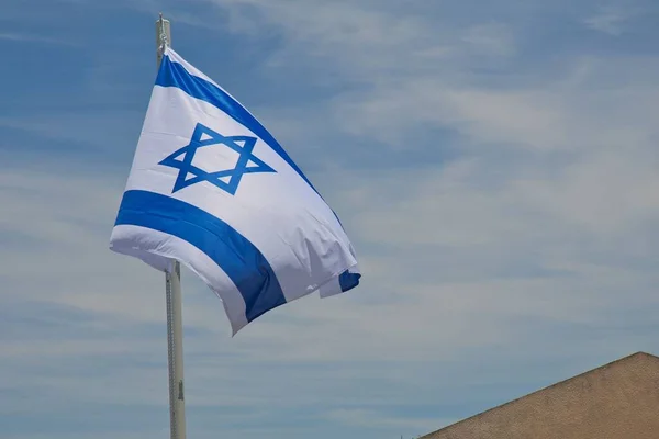 The flag of Israel on pole flutters in wind under blue sky. Israeli flag with the star of David