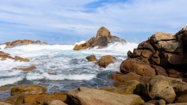 Güneşli bir günde kumlu bir sahilde kayalara çarpan okyanus dalgalarının manzarası.