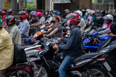 Motosikletli bir kadın kask takmıyor, motosikletliler kırmızı ışıkta bekliyor.