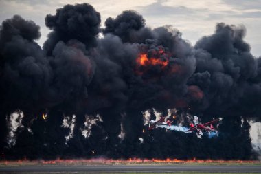 Tarlada siyah dumanlı büyük bir yangın ve ateşin yanında uçan üç uçak.