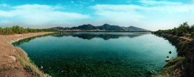 Arka planda dağlar olan bir gölün panorama çekimi. Mavi gökyüzü ve suyun yansıması