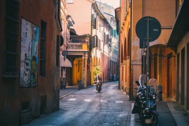 İtalya, Bolonya 'da gündüz vakti renkli binaları olan küçük dar bir yolda scooter süren bir adam.