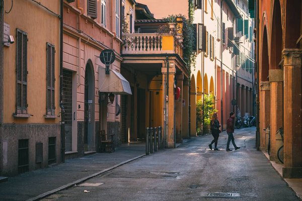 A two people crossing a small road with colorful buildings in Bologna, Italy on sunny day