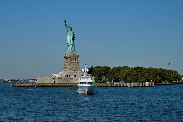 A white yacht moving towards the camera in New York Bay with the Statue of Liberty in the background
