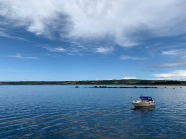 Sakin sularda, mavi bulutlu gökyüzünün altında dağları olan küçük beyaz bir gemi.